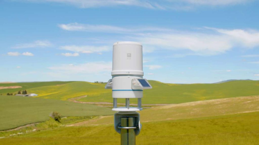 A photograph of an ATMOS 41W all-in-one weather station mounted on a pole with rolling hills of fields in the background