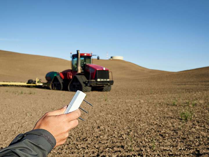 A photograph of a researcher holding a TEROS 12 soil moisture sensor in the foreground and a bare field with a plow in the background