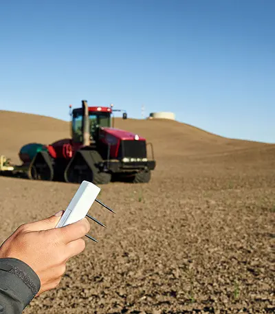 A photograph of a researcher holding a TEROS 12 soil moisture sensor in the foreground and a bare field with a plow in the background